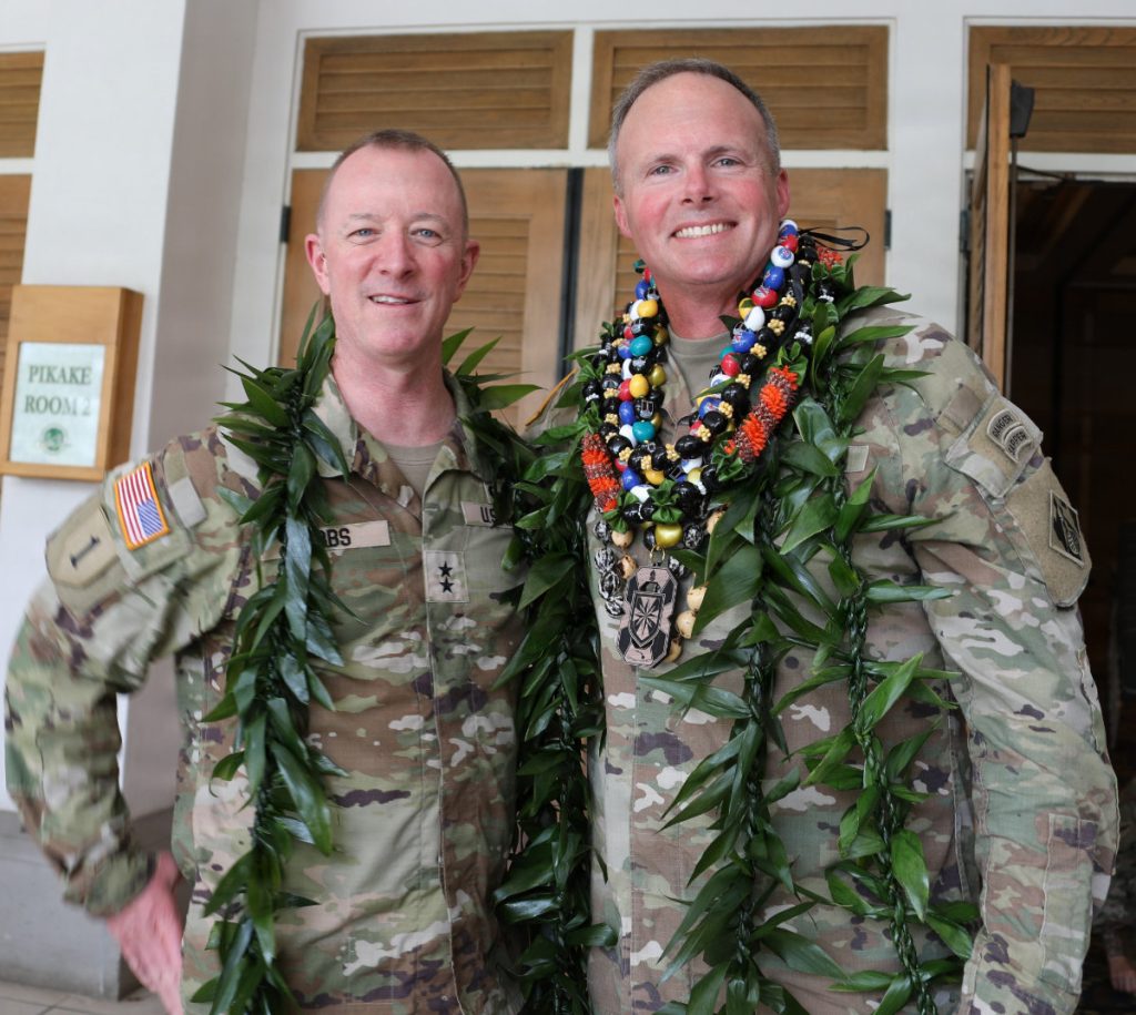 Two U.S. Army officers in camouflage operational uniforms smiling together while wearing traditional Hawaiian leaf and nut leis.