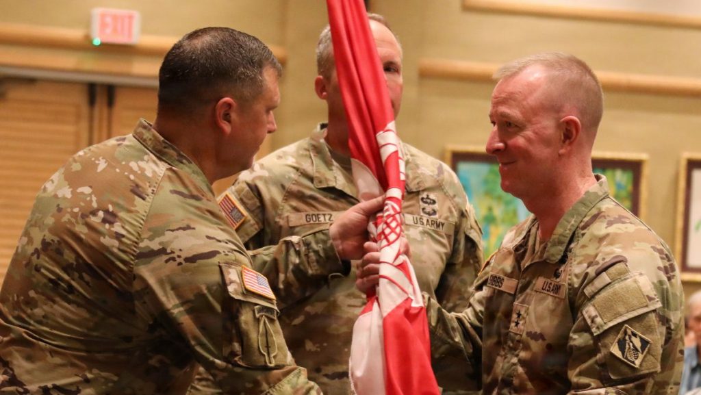 Close-up of military leaders holding and passing the red organizational colors inside a decorated event room.
