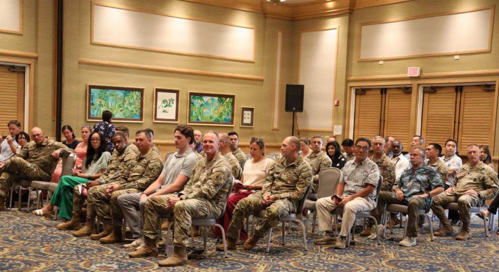 Military personnel and civilian guests seated in rows inside a hall watching an official ceremony.