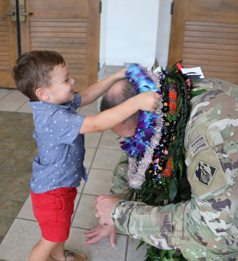 A young boy in a blue shirt and red shorts places a purple flower lei around the neck of an Army officer kneeling before him.