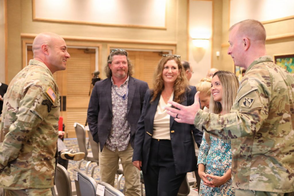 A general officer gestures while speaking with fellow soldiers and civilian guests during an indoor post-ceremony reception.