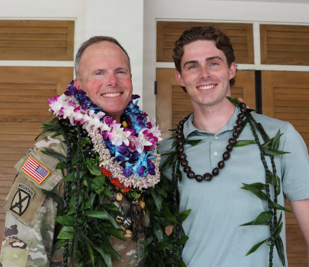 An Army officer heavily adorned with Hawaiian flower and kukui nut leis smiles alongside a young adult family member wearing a leaf lei.