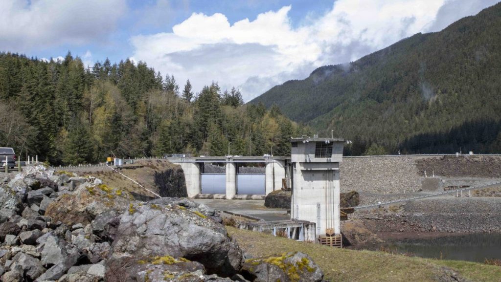 A concrete intake tower and spillway gates at the Howard A. Hanson Dam surrounded by rocky terrain and forested mountain slopes under a partly cloudy sky.