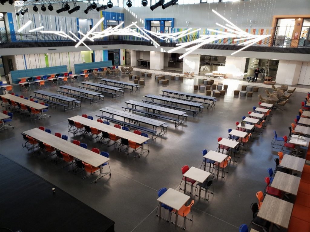 An aerial view of a large multi-level indoor cafeteria filled with rows of dining tables, colorful chairs, and a modern zigzag overhead light fixture.