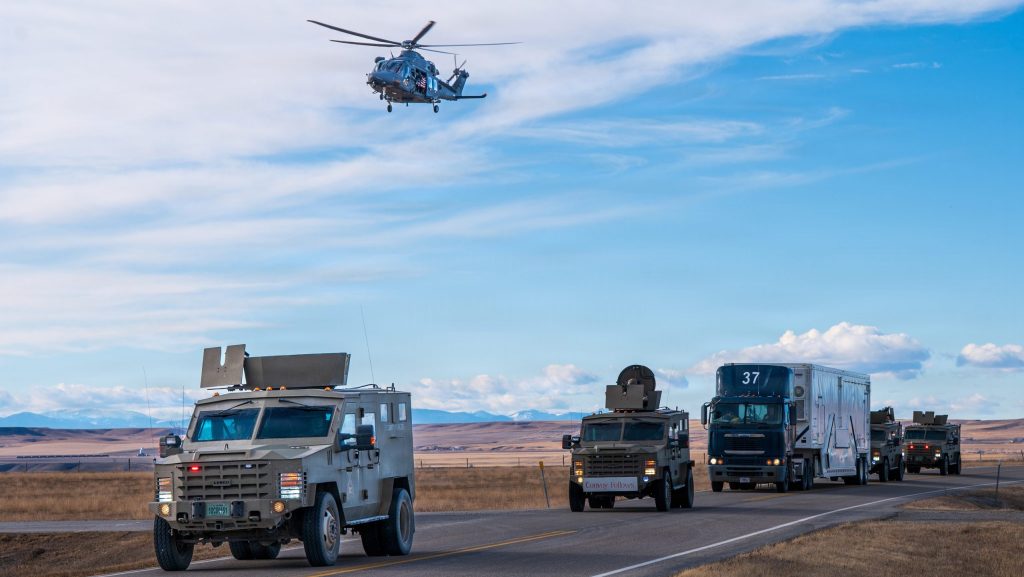 An MH-139A Grey Wolf helicopter flies low over an armored tactical security convoy escorting a transport trailer down a highway.
