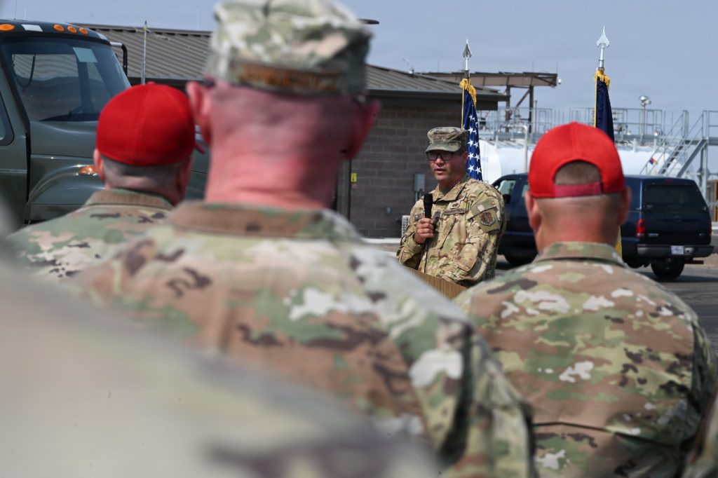 An Air National Guard officer speaks at a podium as seen over the shoulders of service members wearing red baseball caps.