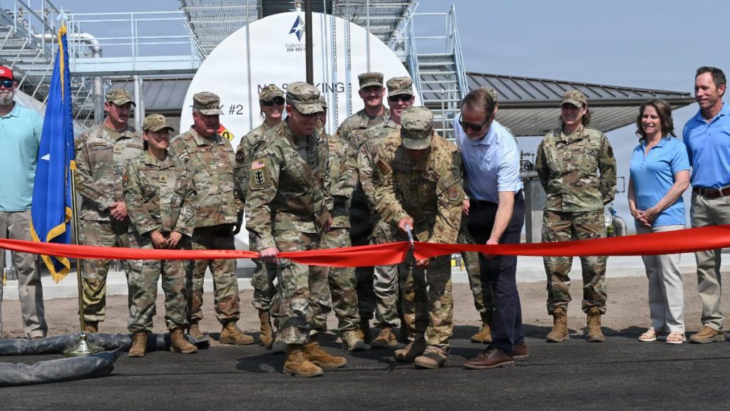 Military officers and civilian representatives cut a bright red ribbon with large ceremonial scissors in front of a new fuel storage installation.