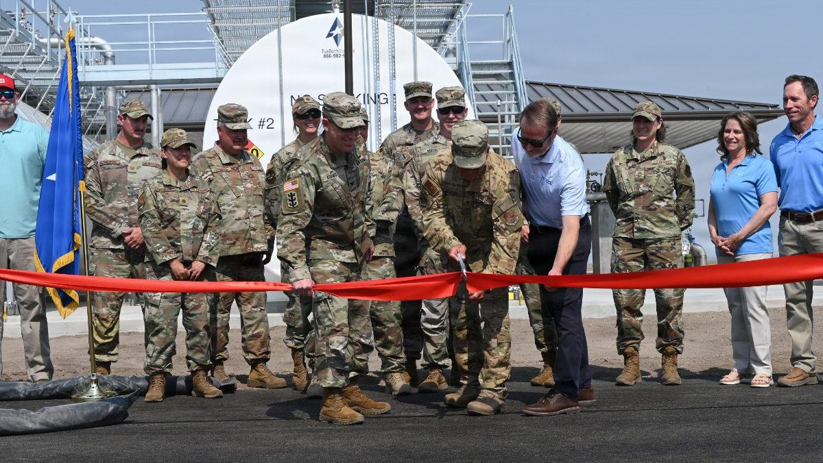 Military officers and civilian representatives cut a bright red ribbon with large ceremonial scissors in front of a new fuel storage installation.