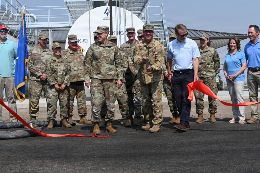 Military commanders and project leaders cheer and walk past a freshly cut red ribbon during an outdoor ribbon-cutting ceremony.