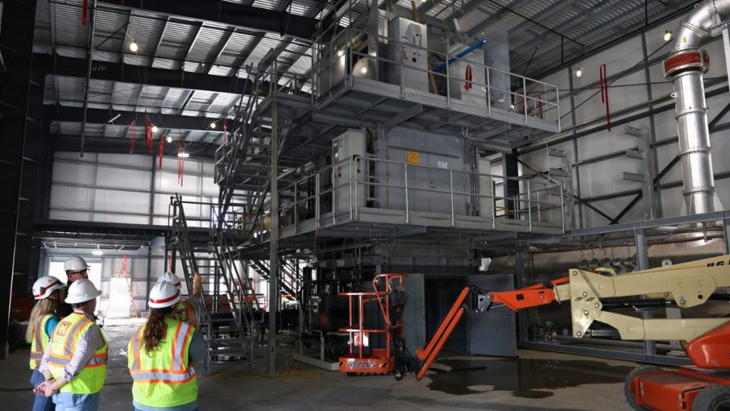 Engineers and site inspectors in safety gear look up at a multi-story industrial processing frame and boom lift inside a large warehouse facility.