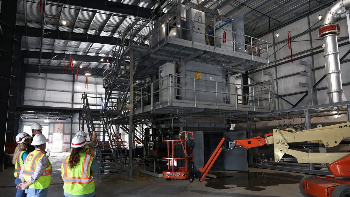 Engineers and site inspectors in safety gear look up at a multi-story industrial processing frame and boom lift inside a large warehouse facility.
