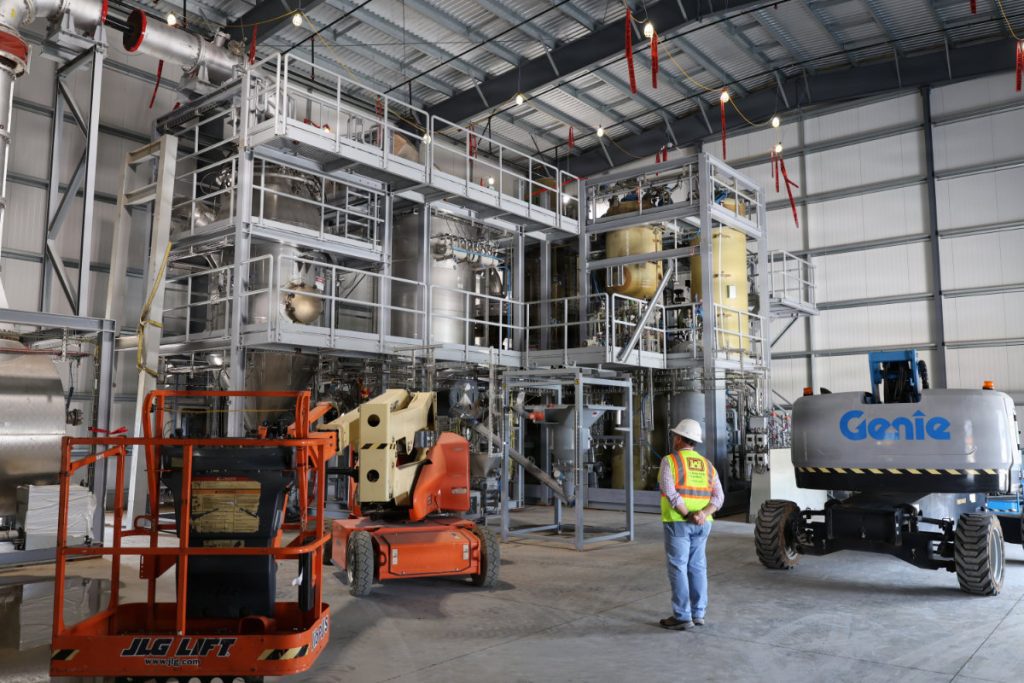 An inspector in a high-visibility vest stands inside a high-bay facility viewing a complex multi-level steel structure, piping, and lifts.