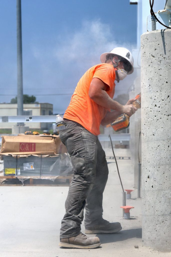 A construction worker wearing a white hard hat, face mask, and bright orange shirt uses a power tool near a concrete support column on an outdoor site.