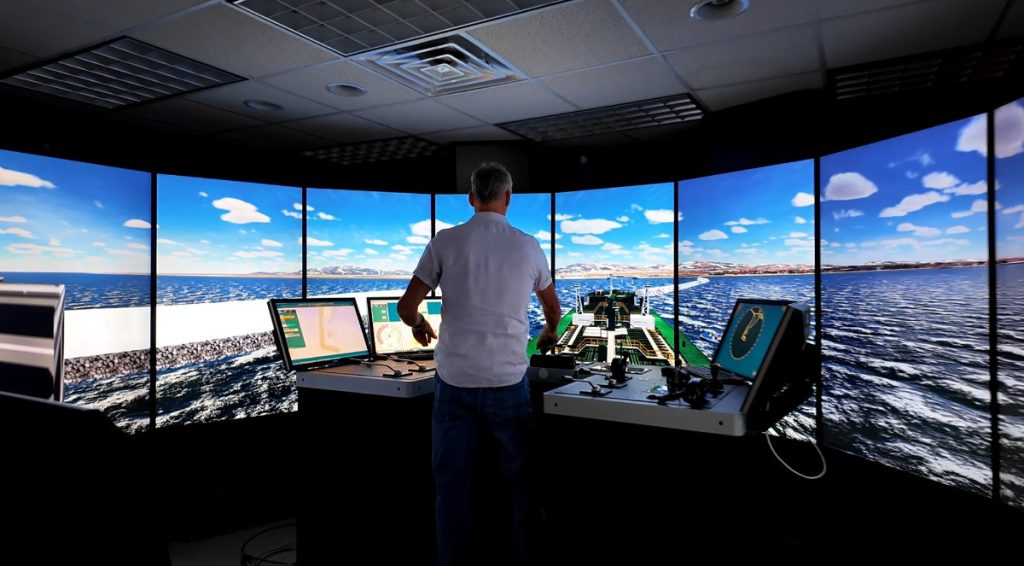 A man stands at a ship navigation simulator console surrounded by panoramic curved screens displaying a 3D vessel bow navigating open water.