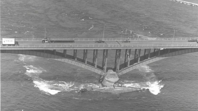 An aerial black-and-white photograph of a partially submerged vessel lodged against a concrete bridge pier in turbulent river water with traffic crossing above.