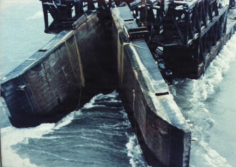 An overhead view of a partially submerged split-hull dump barge opening in choppy water during emergency operations.