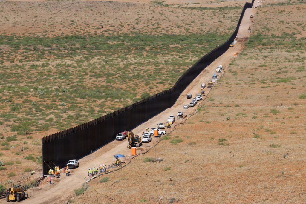 An aerial view of a tall bollard barrier wall stretching across desert terrain with construction vehicles and trucks parked along the access road.