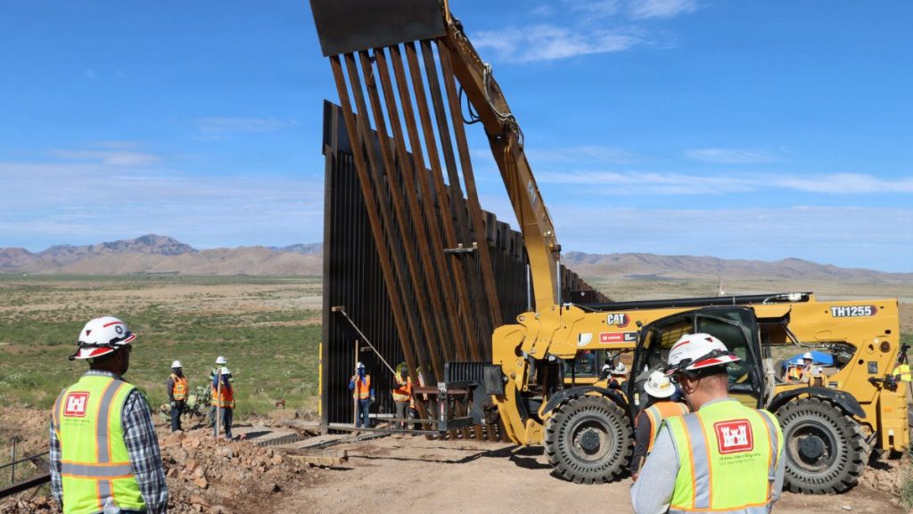 A heavy CAT telehandler positions a steel bollard panel vertically while USACE engineers in safety gear monitor the installation from the foreground.