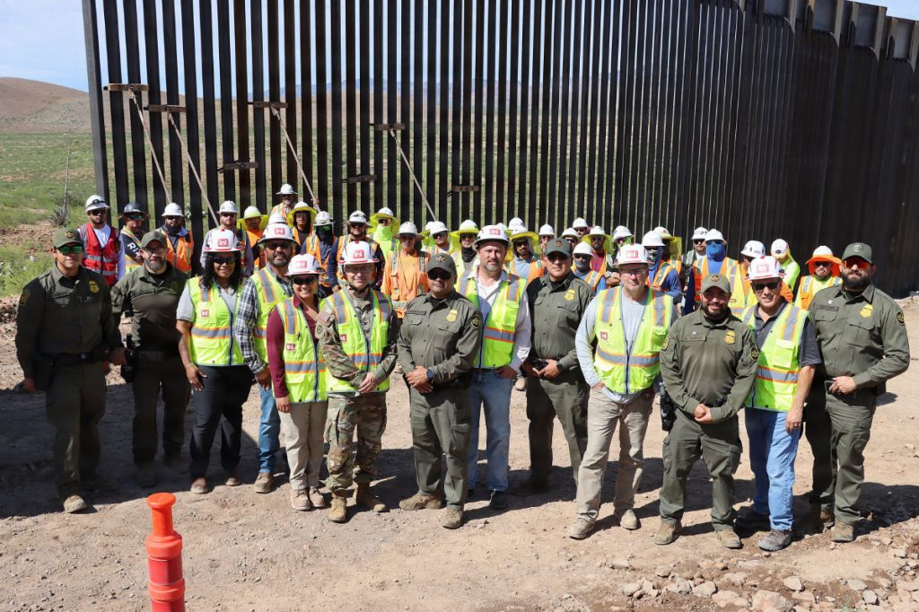 A large group of construction workers, USACE officials, and Border Patrol agents pose for a group photo in front of a tall metal border wall structure.