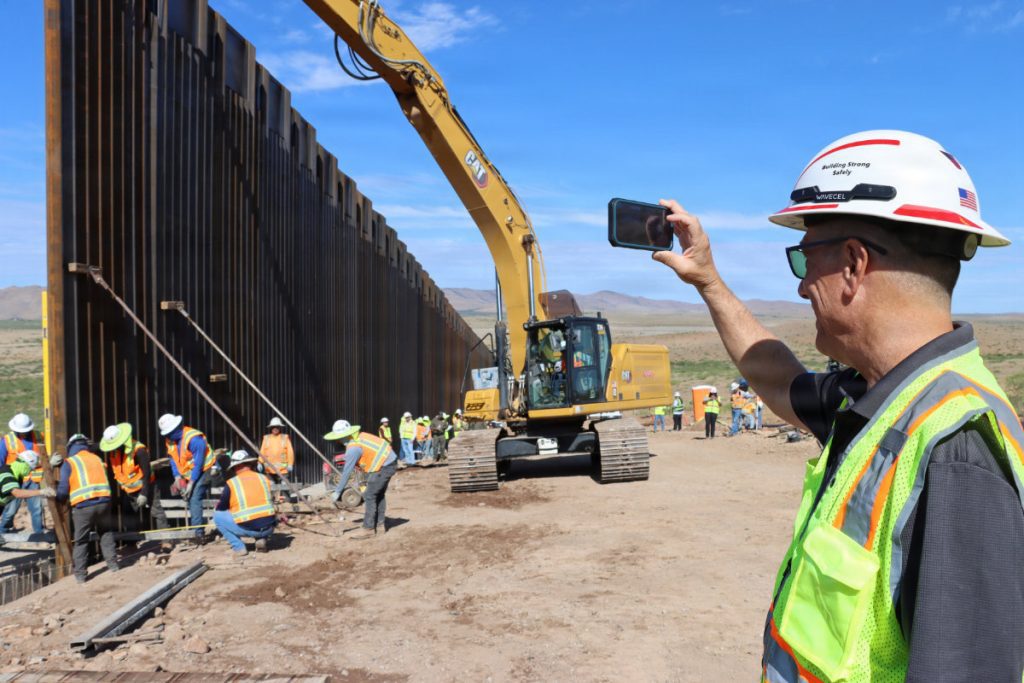 A safety official in a hard hat takes a smartphone photo of ground crews securing a metal wall panel lifted by a yellow CAT excavator.