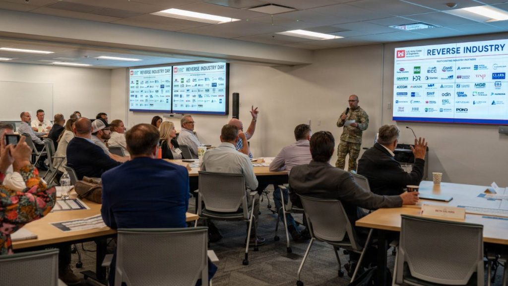 A military officer speaks into a microphone during a conference presentation in a room filled with seated industry partners raising their hands, with screens displaying "Reverse Industry Day" partner logos.
