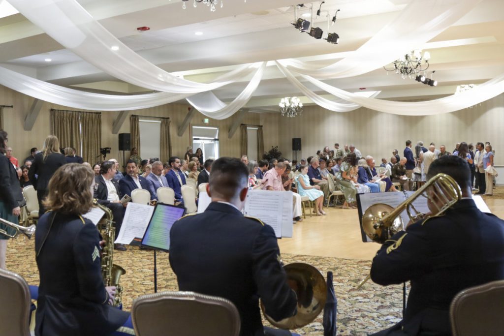 A military band plays brass and woodwind instruments in the foreground of a large banquet hall filled with seated guests under white ceiling drapes.
