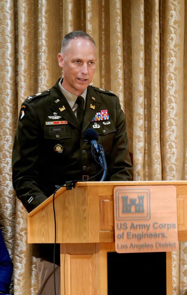 A U.S. Army colonel in dress uniform speaks into a microphone at a wooden lectern bearing the USACE Los Angeles District emblem.