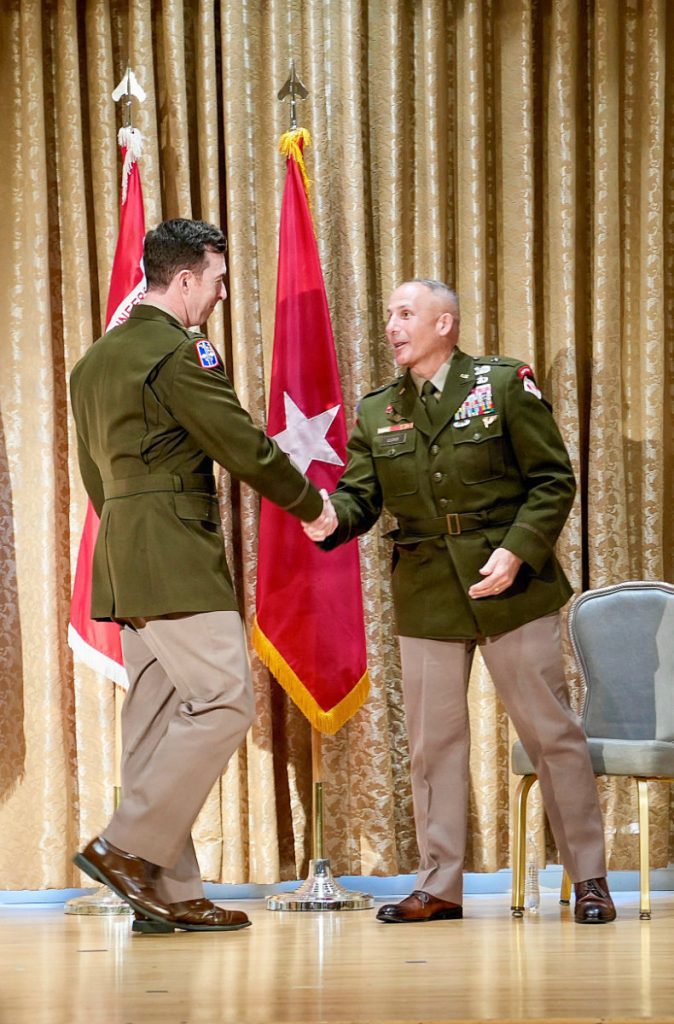 Col. Joshua Bost handshakes a general officer on stage in front of military flags during a formal indoor military event.