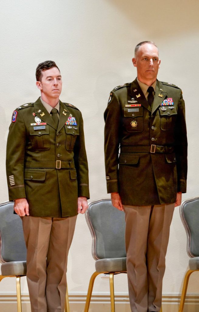 Col. Joshua Bost and a fellow Army officer stand at attention side-by-side in dress uniforms in front of stage seating.