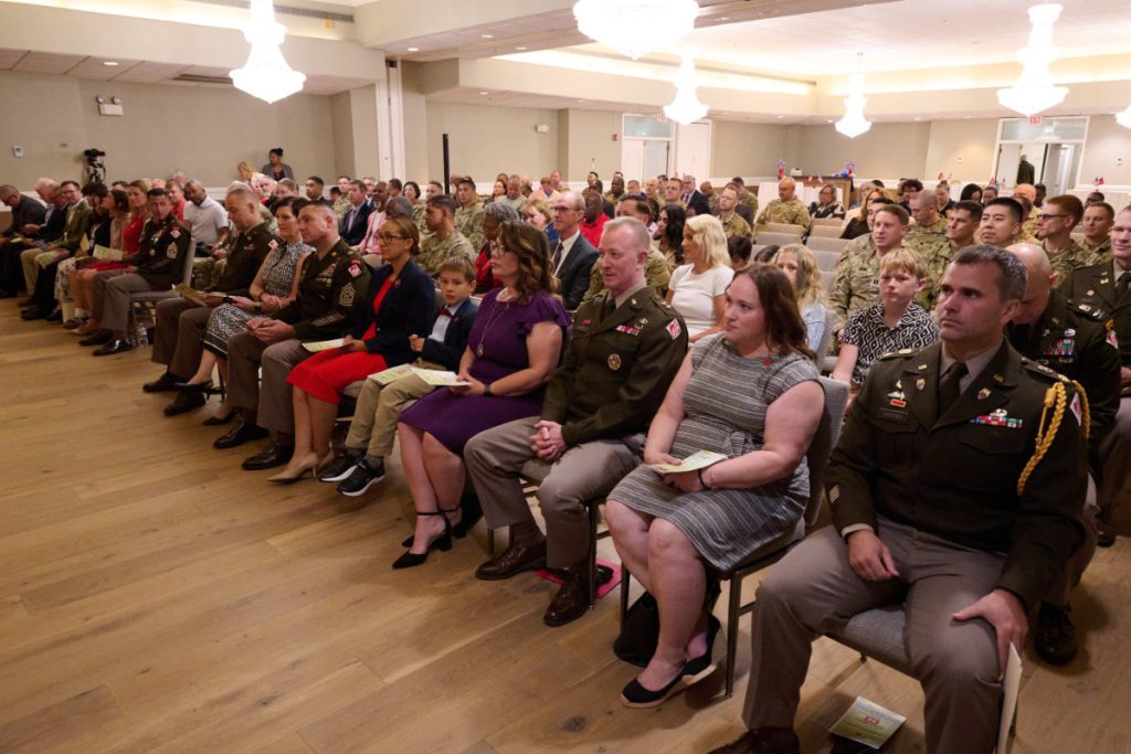 Distinguished guests, family members, and military personnel in Army Green Service Uniforms sit in rows during an indoor ceremony inside a banquet hall.