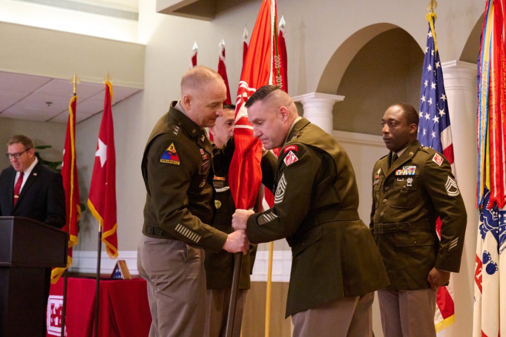 Close-up of a three-star general handing the red unit flag to the incoming command sergeant major standing beside other senior Army NCOs.