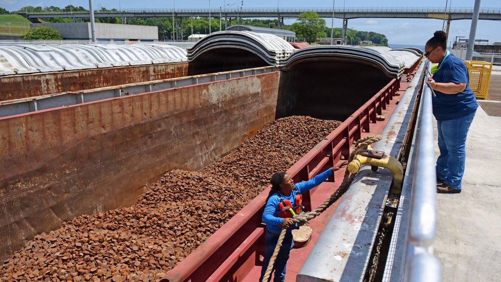 A crew member aboard a cargo barge filled with gravel secures a thick mooring rope to a lock wall fixture while a lock operator watches from above.