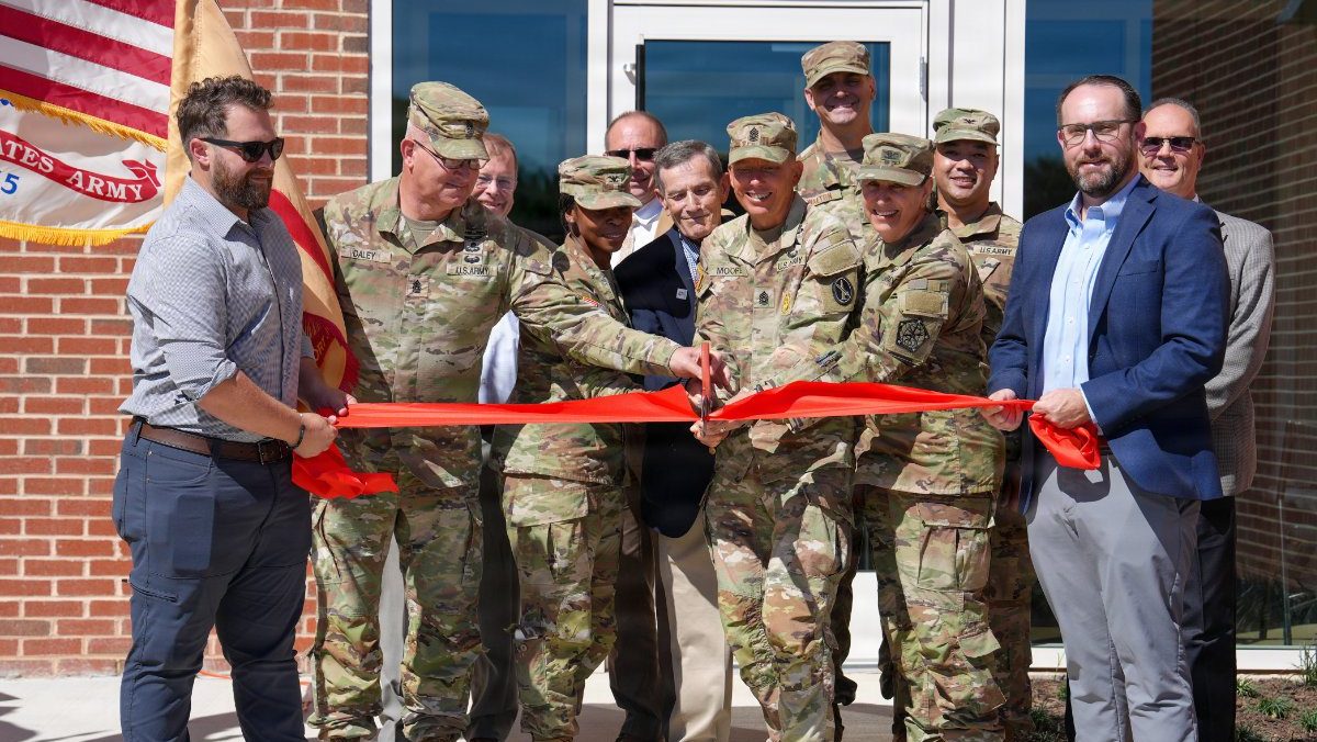 Military leaders and civilian officials gather in front of a brick building to cut a wide red ribbon together using oversized scissors.