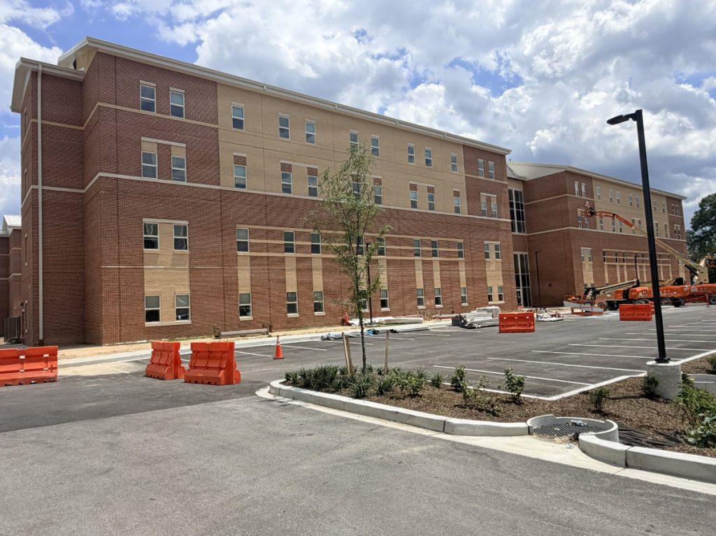 Exterior view of a multi-story red brick barracks facility featuring a paved parking lot, young landscaping trees, and orange safety barricades.