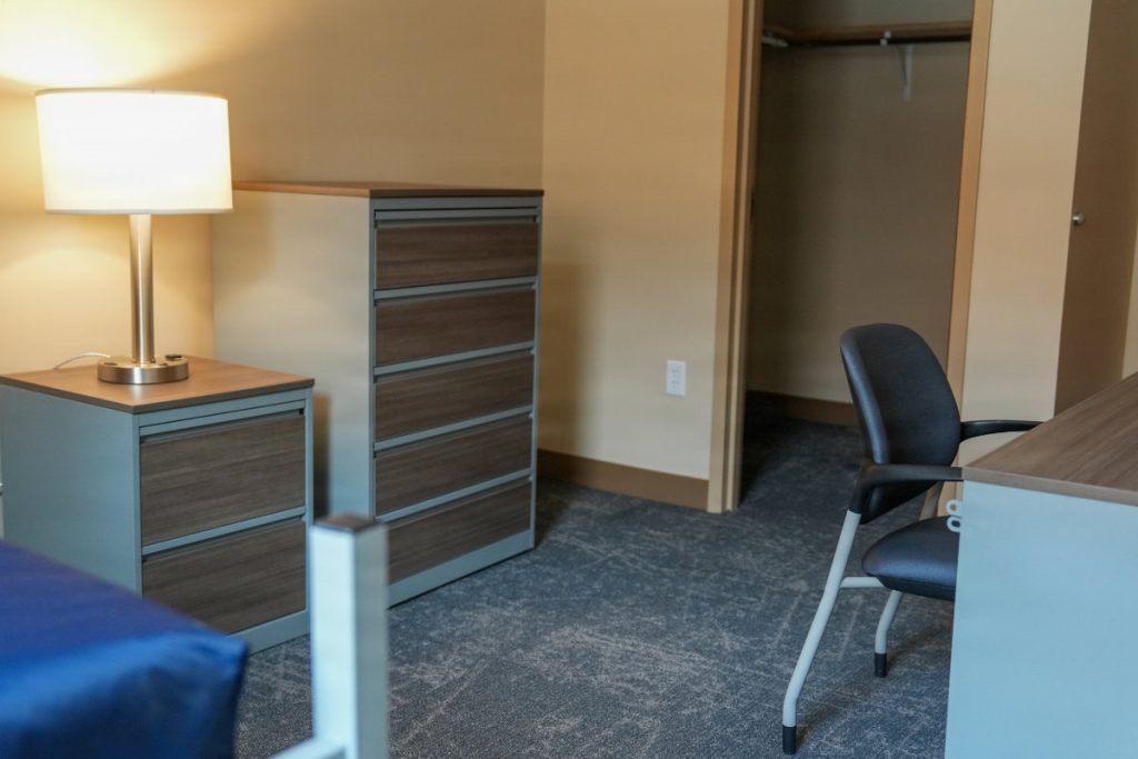 A military barracks bedroom layout displaying a desk with an office chair, a nightstand with a lamp, a chest of drawers, and an open closet doorway.