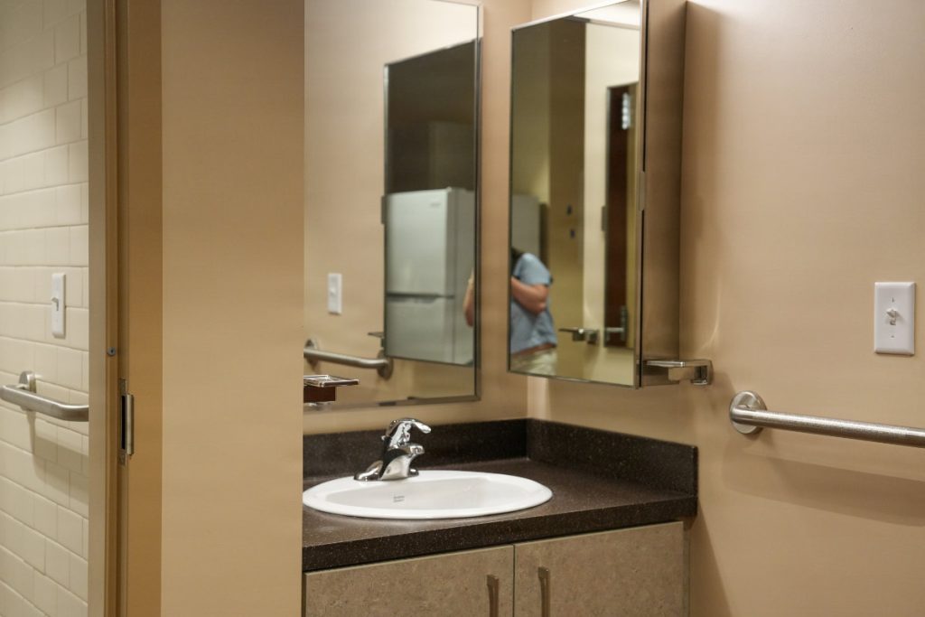 A modern bathroom interior featuring a clean white sink basin, dark countertop, medicine cabinet mirror, and stainless steel grab bars.