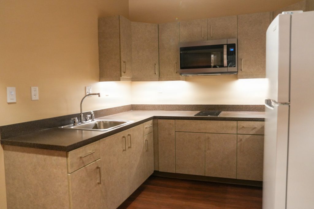 A compact kitchenette in a service member's room with tan cabinetry, a stainless steel sink, built-in microwave, cooktop, and white refrigerator.