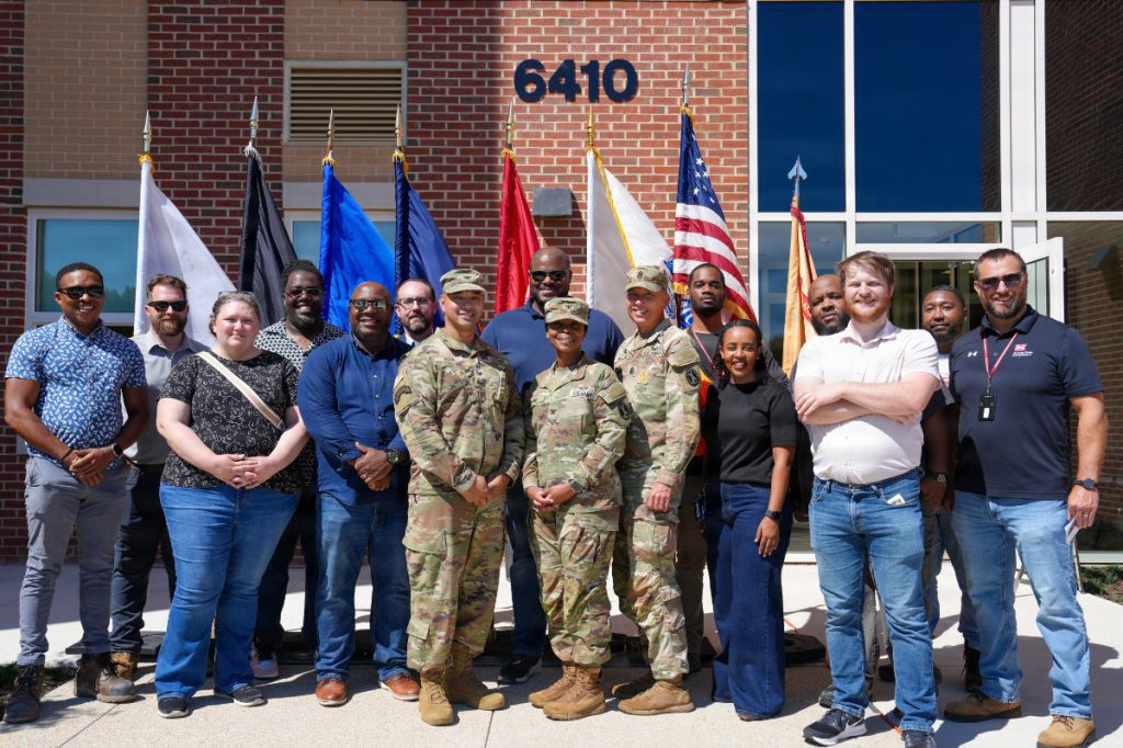 A group of military personnel and civilian team members pose for a group photo in front of service flags and a brick building entrance numbered 6410.