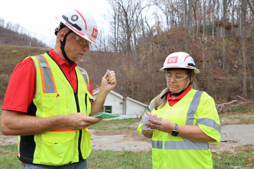Two safety officials in hard hats, safety glasses, and high-visibility vests taking notes on small pads outdoors near a wooded hillside.