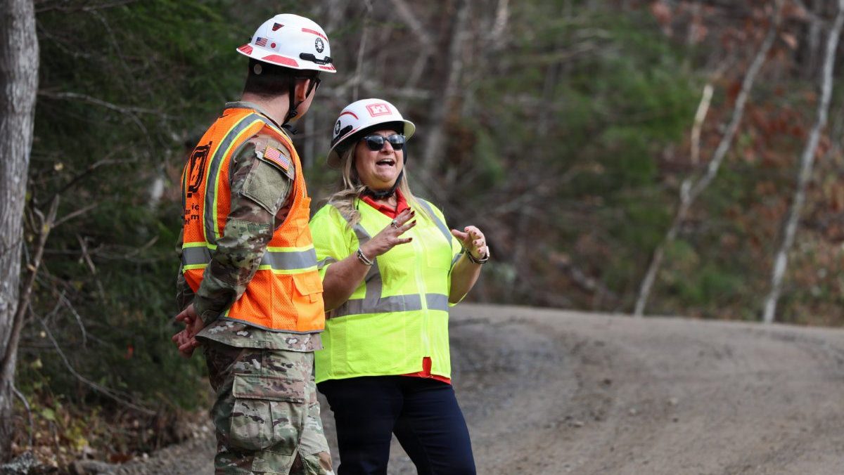 A woman emergency manager in a high-visibility vest gestures while speaking with an Army soldier in camouflage along a dirt road.