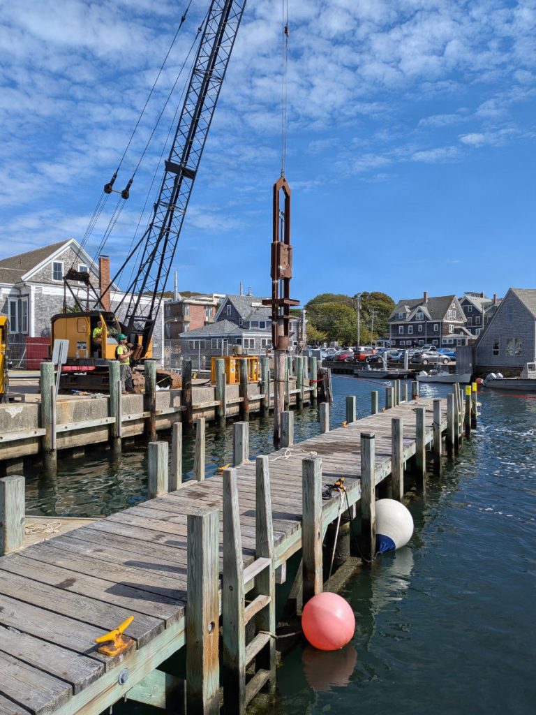 A yellow construction crane operates an impact hammer to drive a timber pile into the water alongside a wooden pier in a coastal harbor.