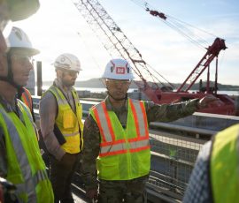 Close-up profile of a USACE officer in an OCP uniform and "Building Strong" hard hat gesturing with his hand while speaking to a group of personnel on a construction site.