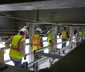 Four workers in safety vests walking away from the camera through a narrow, industrial gray metal corridor located underneath a bridge or large concrete structure.